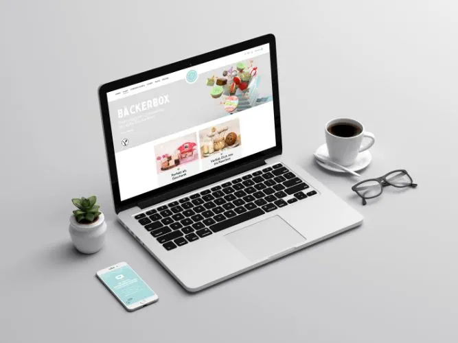 Startseite von BäckerBox an einem Laptop, mit einer Tasse, einer Brille, einer Pflanzen und einem Handy daneben